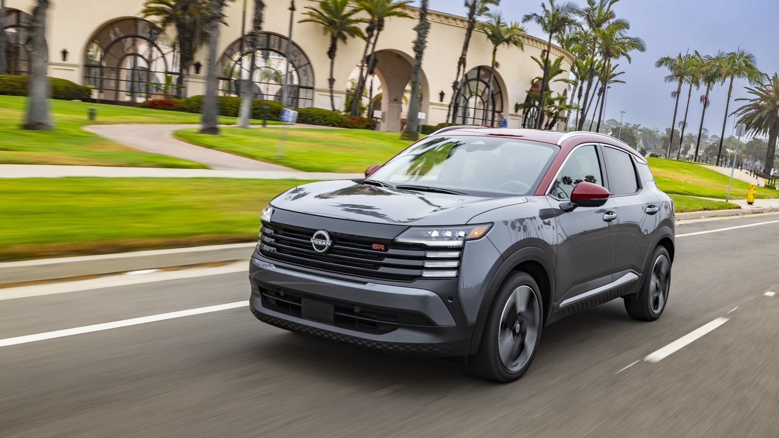 2026 Nissan Kicks SR in Gun Metallic with red contrast roof, front three-quarter motion shot on a palm-lined urban boulevard